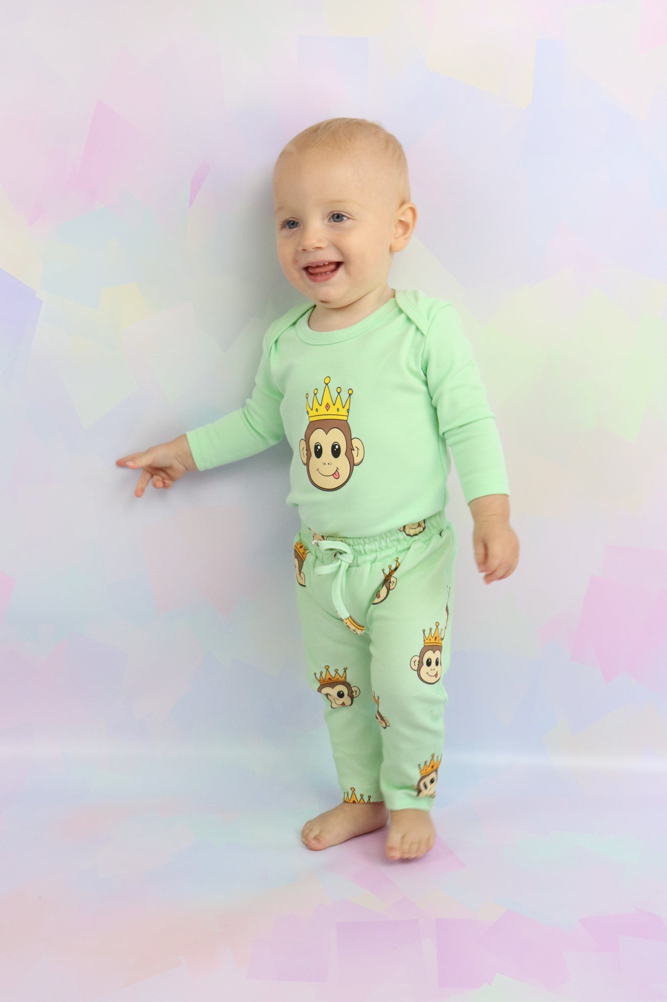 toddler boy standing up and smiling, wearing a pastel green long sleeve romper with a cute monkey design on it. Also wearing pastel green pants with the cute monkey design printed all over