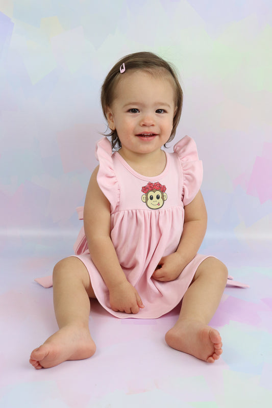 toddler girl sitting down wearing a pastel pink dress with a cute monkey face printed on it. Dress has pretty frill sleeves and tie-up bow detail on the sides