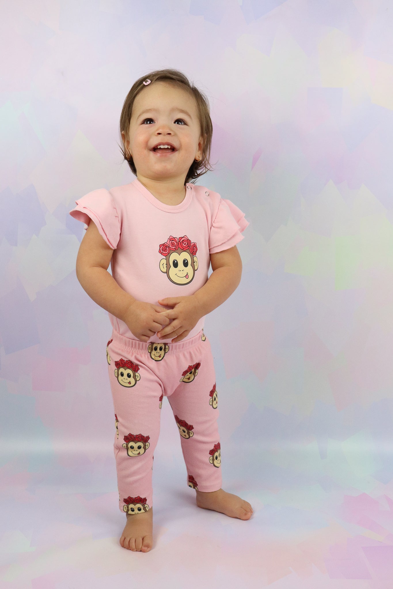toddler girl wearing a frilly pink romper with a cute monkey face printed on it. Also wearing pink leggings with cute monkey faces printed all over