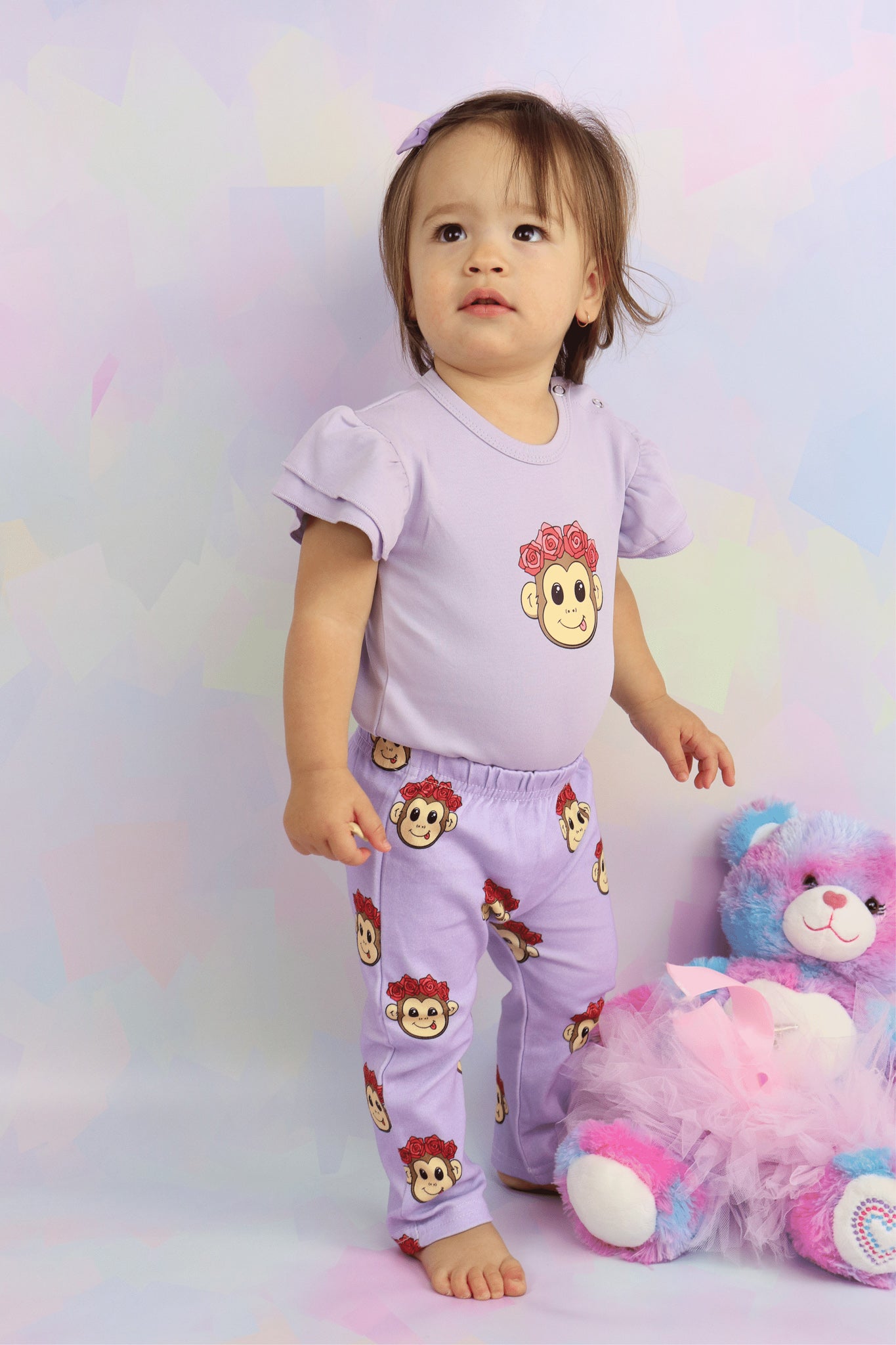 toddler girl standing up, wearing a pastel purple romper with a cute monkey face design on it. Romper has frill short sleeves. Also wearing matching purple pants with cute monkey faces printed all over