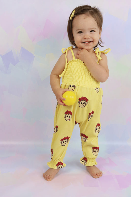 toddler girl wearing a pastel yellow jumpsuit with cute monkey faced printed all over. Shirred top with tie-up straps