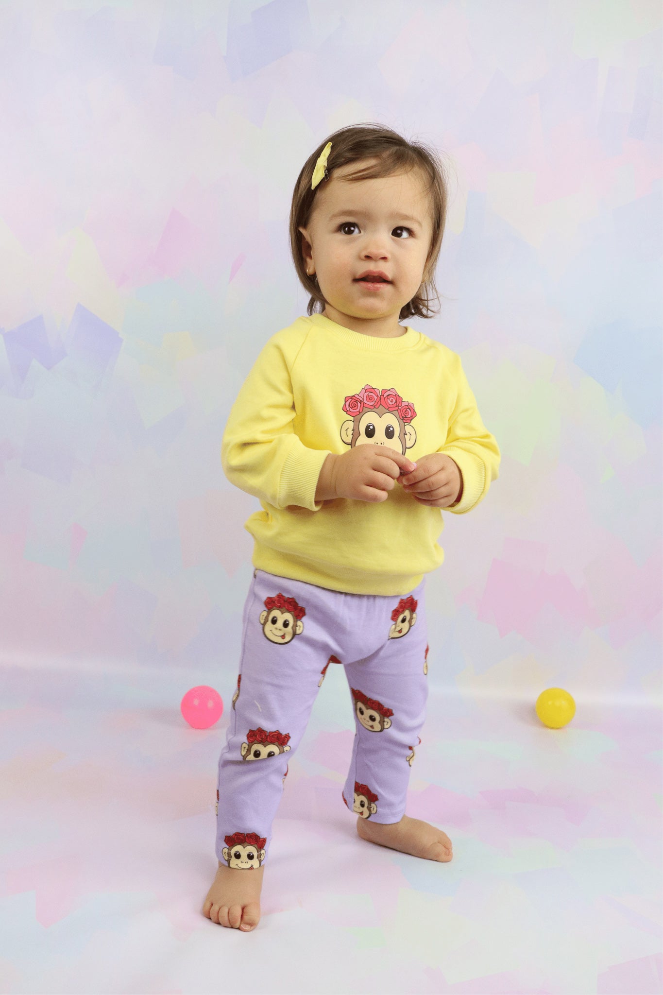 toddler girl wearing a pastel yellow jumper with a cute monkey faced printed on it. Also wearing pastel purple leggings with the cute monkey faced printed all over