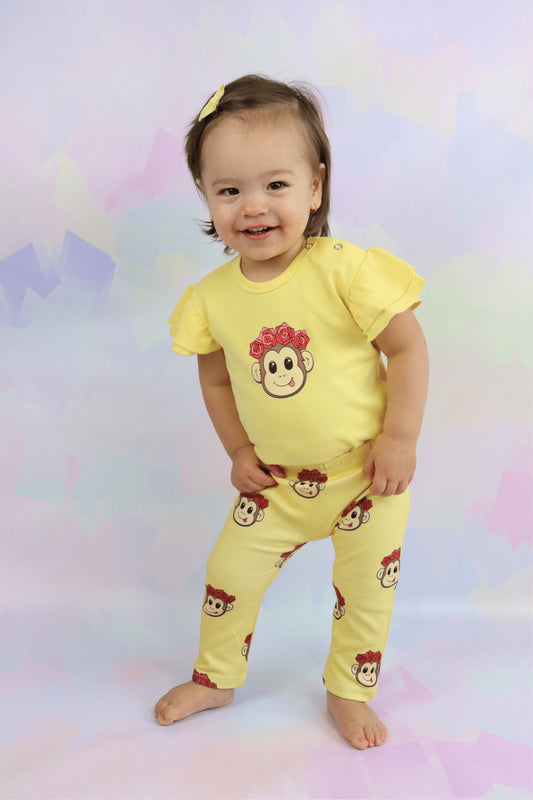 toddler girl standing up wearing a pastel yellow short sleeve frill romper with a cute monkey face on it. Also wearing matching yellow pants with the monkey face printed all over